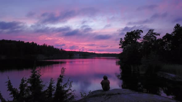 Aerial drone shot,of a man sitting at a rocky shore, of a lake, a purple sky, a colorful sunset or d alt