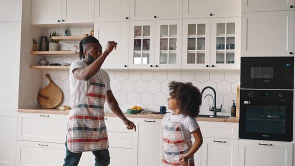 Happy African American Father Dancing with His Little Daughter at Kitchen Wearing Aprons Giving High alt