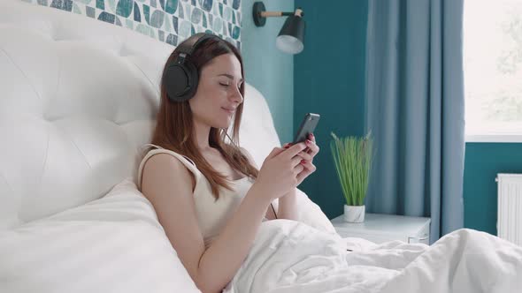 Relaxed Young Brunette Woman in Headphone Lying on White Bed with Smartphone in Hands alt