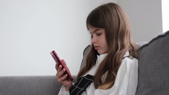 a Small Young Beautiful Girl in a School Uniform Sits on a Sofa with a Phone in Her Hands