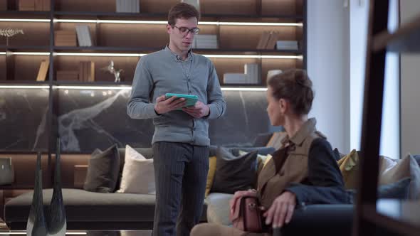 Wide Shot Male Manager Consulting Female Client Sitting on Soft Couch in Furniture Showroom alt