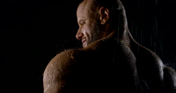 Naked Male Culturist Is Standing Under Water Flows in Darkness, Back View on Big Shoulders alt