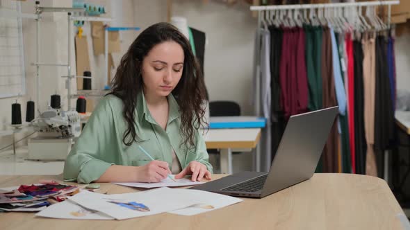 Focused young female tailor writing notes in paper, using laptop. Concentrated fashion designer alt