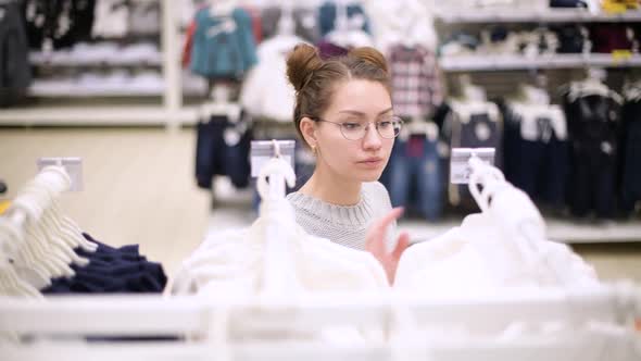 a young mother walks along a row of children's clothes, choosing a sweater for her child alt
