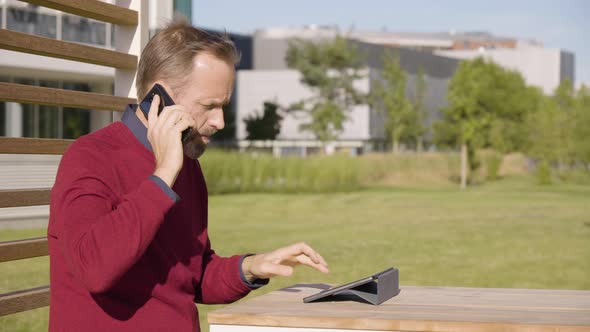 A Middleaged Handsome Caucasian Man Talks on a Smartphone and Works on a Tablet As He Sits alt