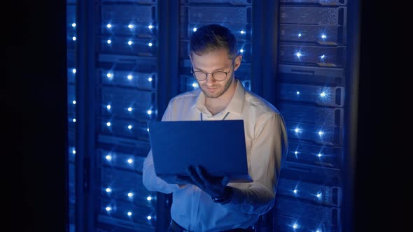 Male Network Engineer Doing a System Check Standing in the Server Room with His Laptop alt