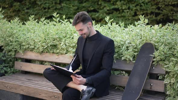 Joyful Happy Young Businessman Looking Through Paperwork Sitting on Bench in City Park and Leaving alt