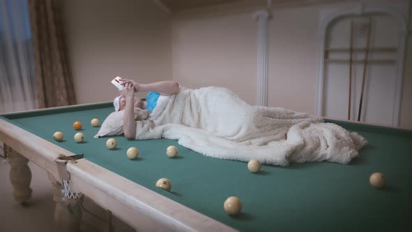 Man with a cell phone in his hands lies on a pool table, covered with a blanket alt