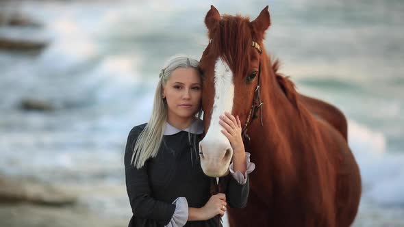 Beautiful Girl in a Long Dress is Standing and Hugging a Bay Horse on the Beach