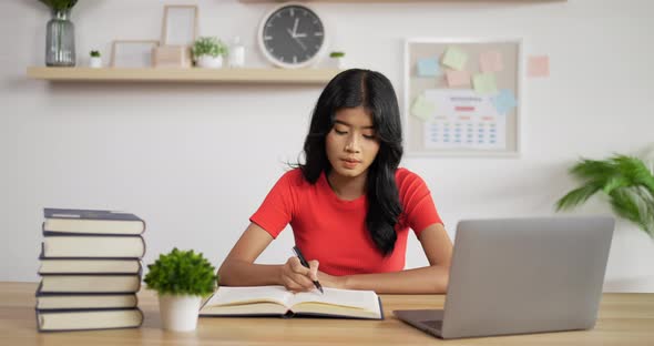Portrait of Asian schoolgirl studying online making notes in copybook alt