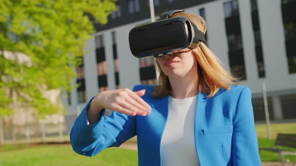 A Happy Woman in the Suit is Standing Outside and Gestures in Augmented Reality in 3D VR Goggles alt