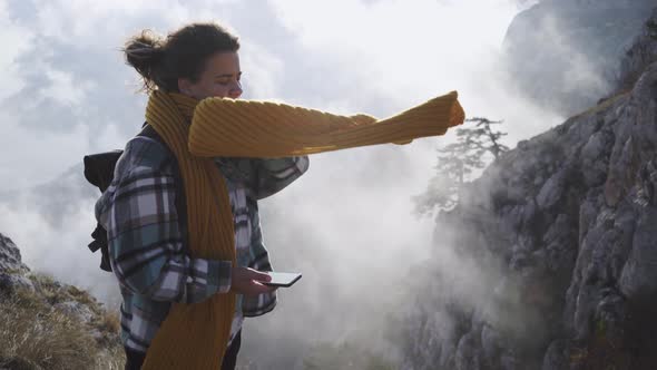 Woman Tourist with Smartphone Adjusts Scarf on Steep Cliff