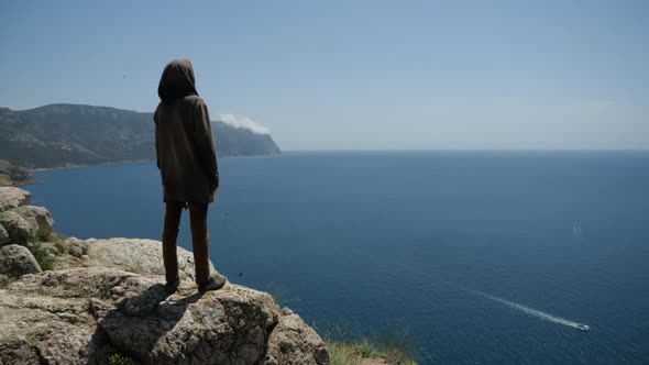 A Man in a Hood on the Edge of the Cliff in Front of Him Has a View of the Sea