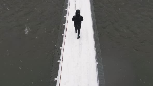 Top Down Shot Man in Black Jacket Makes Drone Footage Himself and Walks Across a Snowcovered Bridge