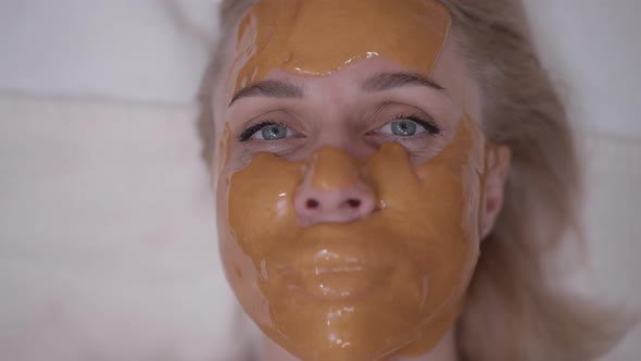 Closeup Slim Blond Caucasian Woman in Yellow Facial Mask Looking at Camera with Grey Eyes Smiling alt