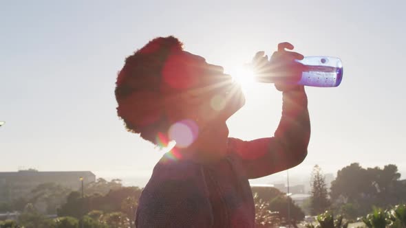 Fit african american man exercising outdoors in city, resting and drinking from water bottle alt