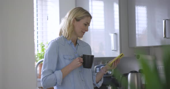 Blond woman with coffee cup standing in kitchen reading text messages