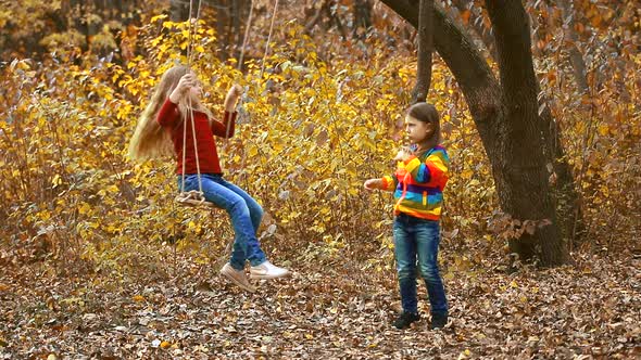 Two little girls play with rope swing and swing each other in autumn Park. Children laugh happily.