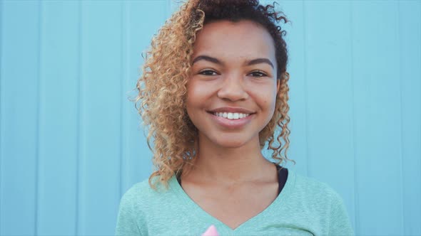 Young Afro Girl Eating Pink Melting Ice Cream at Blue Wall Background alt