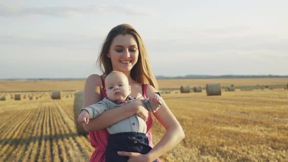 Portrait of Happy Mother Lulls Her Lovely Baby in Hands with Smile in Hay Field alt