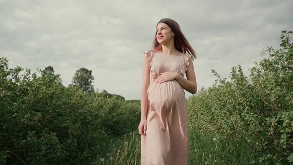 A young pregnant woman with flowing hair in a pink summer dress walks among rows of green plants.