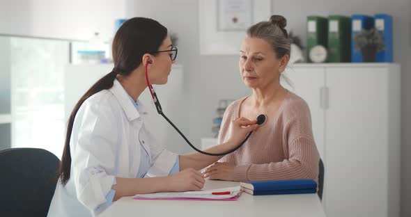 Young Female Doctor in White Coat Listening with Stethoscope to Aged Patient in Office alt