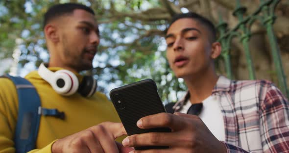 Two happy mixed race male friends standing, talking and using smartphone in the street alt