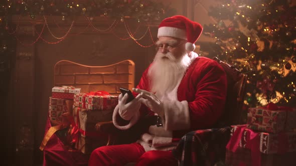 Cute and Funny Santa Claus Is Using Modern Smartphone with Internet, Surfing Social Nets, Portrait alt