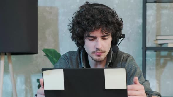 Young Curly Haired Man Using Laptop Computer with Headset to Talking Online alt