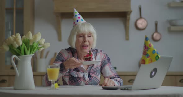 Senior Woman Celebrates Birthday Online with Piece of Cake and Candle. 70s Woman in Birthday Hat alt