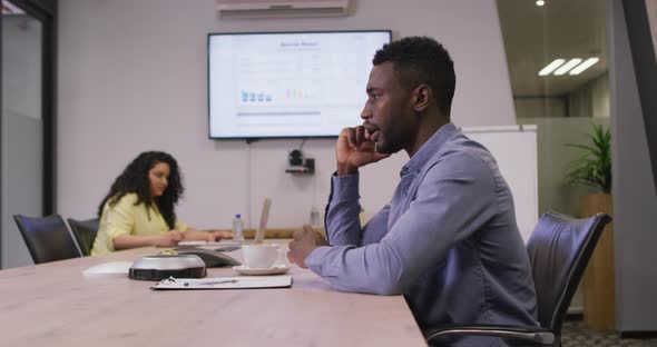 African american businessman sitting at desk, talking on smartphone in modern office alt