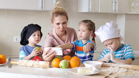 Young Woman Cooks in the Kitchen and Watching the Recipe on Mobile Phone. Children Help Mom To