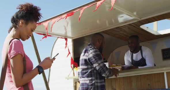 Smiling mixed race woman using smartphone waiting in queue to be served at food truck alt