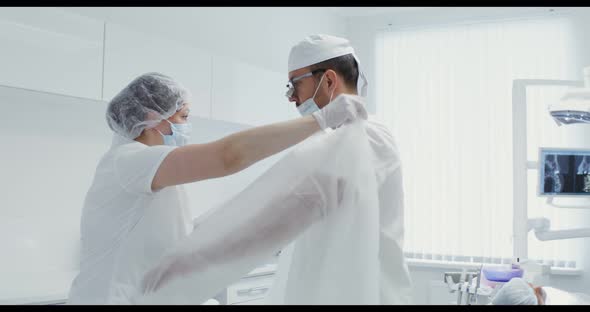 A Nurse Helps a Dentist Put on a White Sterile Suit Tying the Ties of His Back alt