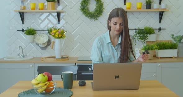 Young Woman Is Making Online Payment Holding Bank Card Using Modern Laptop at Home Sitting on Sofa alt