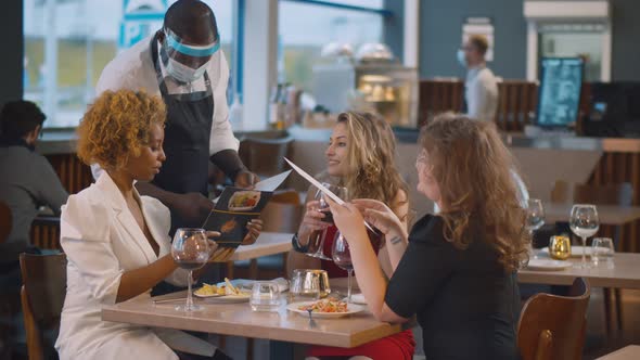 African Waiter in Safety Mask Taking Order From Diverse Group of Customers Sitting at Restaurant alt