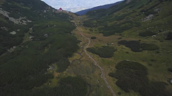 Aerial view of a hiking path in a lush green area, hills left and right and a building in the backgr alt