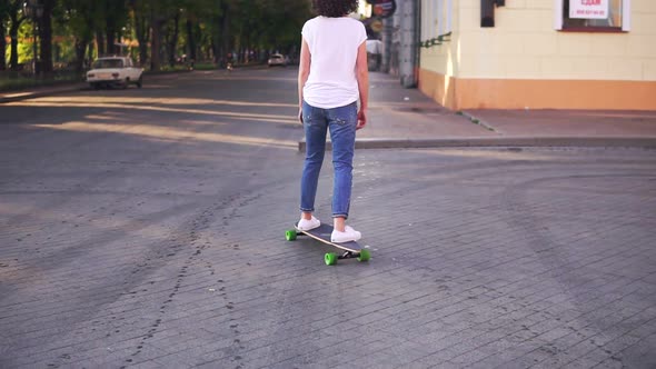 Back View of a Woman in a White Tshirt Blue Jeans and White Sneakers Skateboarding in the City alt