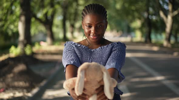 Happy Millennial African American Woman Admiring Toy Rabbit Standing in Sunny Park Outdoors alt