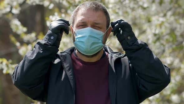 A bearded man in rubber gloves puts on a medical mask against the background of a flowering tree