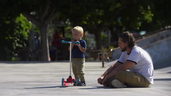 Father with His Little Son in the Skatepark About to Traing Skating on the Scooter alt