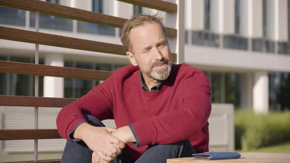 A Middleaged Handsome Caucasian Man Smiles at the Camera As He Sits at a Table in a Park