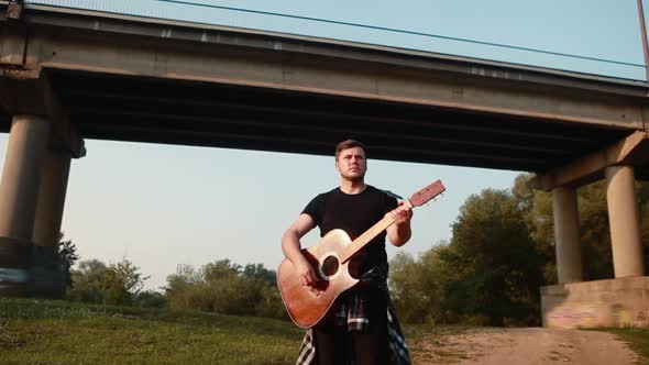 A Young Guy a Guitarist Plays an Acoustic Guitar Under a Bridge with Passing Cars