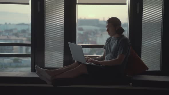 Handsome Hipster Man Sits Alone in Front of a Panoramic Window with a Laptop
