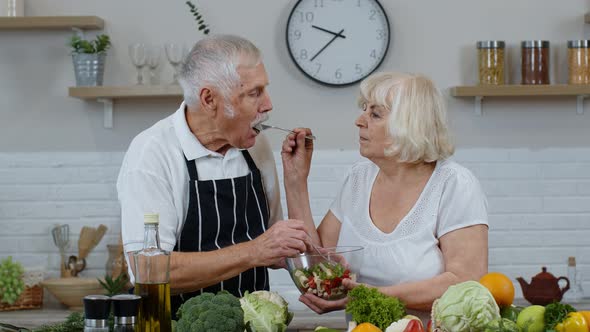 Senior Couple in Kitchen. Grandmother and Grandfather Feeding Each Other with Raw Vegetable Salad alt