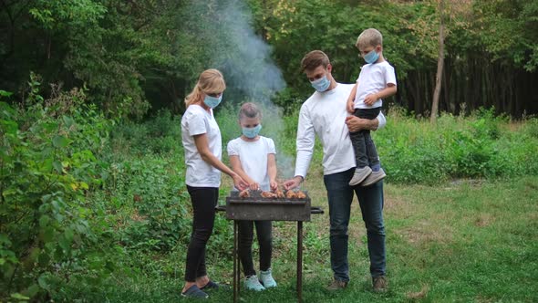 Dad, mom and children fry meat on skewers on the grill. Family in medical masks