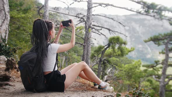 Adventure Trip Young Woman Hiker with Dreadlocks Sitting on the Ground in the Forest and Shooting alt