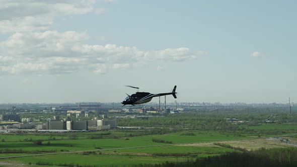 A Dark Helicopter with White Stripes Flies Against a Background of Blue Sky and Green Fields and