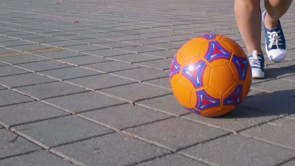 Closeup Boy's Feet Kicking Orange Soccer Ball on Stone Asphalt Surface in City