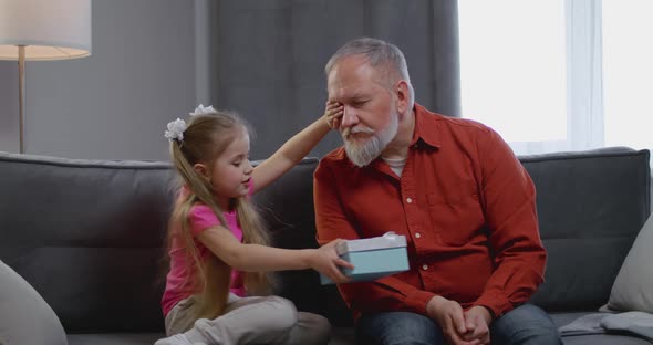 Cute Kid Daughter Make Surprise Present Covering Eyes of Happy Grandpa Receive Gift Box Sit on Sofa